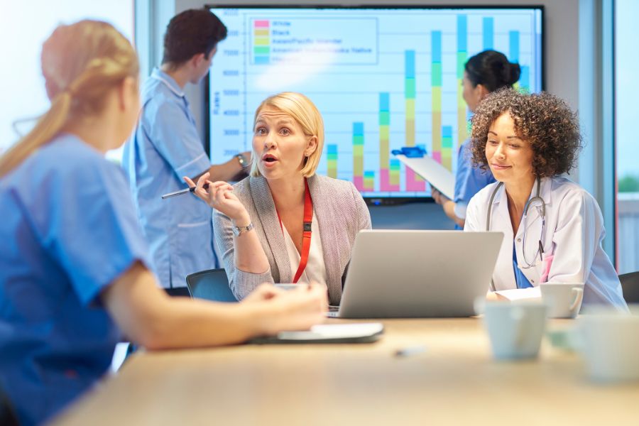 Colaborares de hospital sentados em uma mesa e conversando, gráficos são projetados ao fundo, representando a gestão em saúde